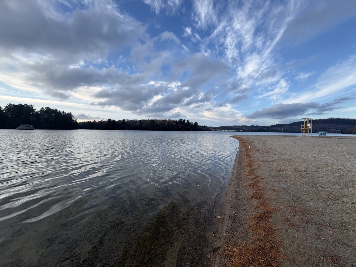 A calm lake meeting a wide, flat sandy beach with a lifeguard tower, a pine-covered far shore and a cloudy sky.