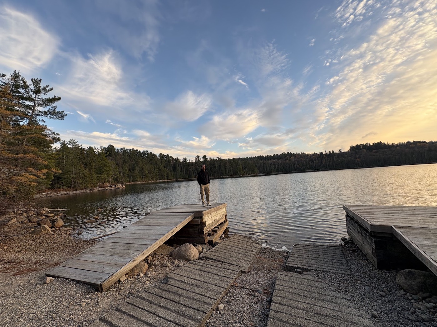 A wooden ramp sloping gently from a firm gravel shoreline down onto a low dock platform beside a calm lake, with a second dock to the right.